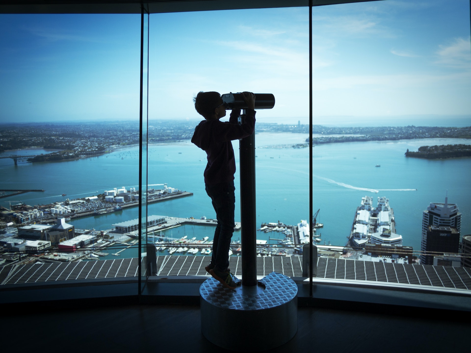 A teen boy steps on tiptoes to see through a telescope inside of the Sky Tower, Auckland, he's almost a siloutte with large ships docked in the waters below